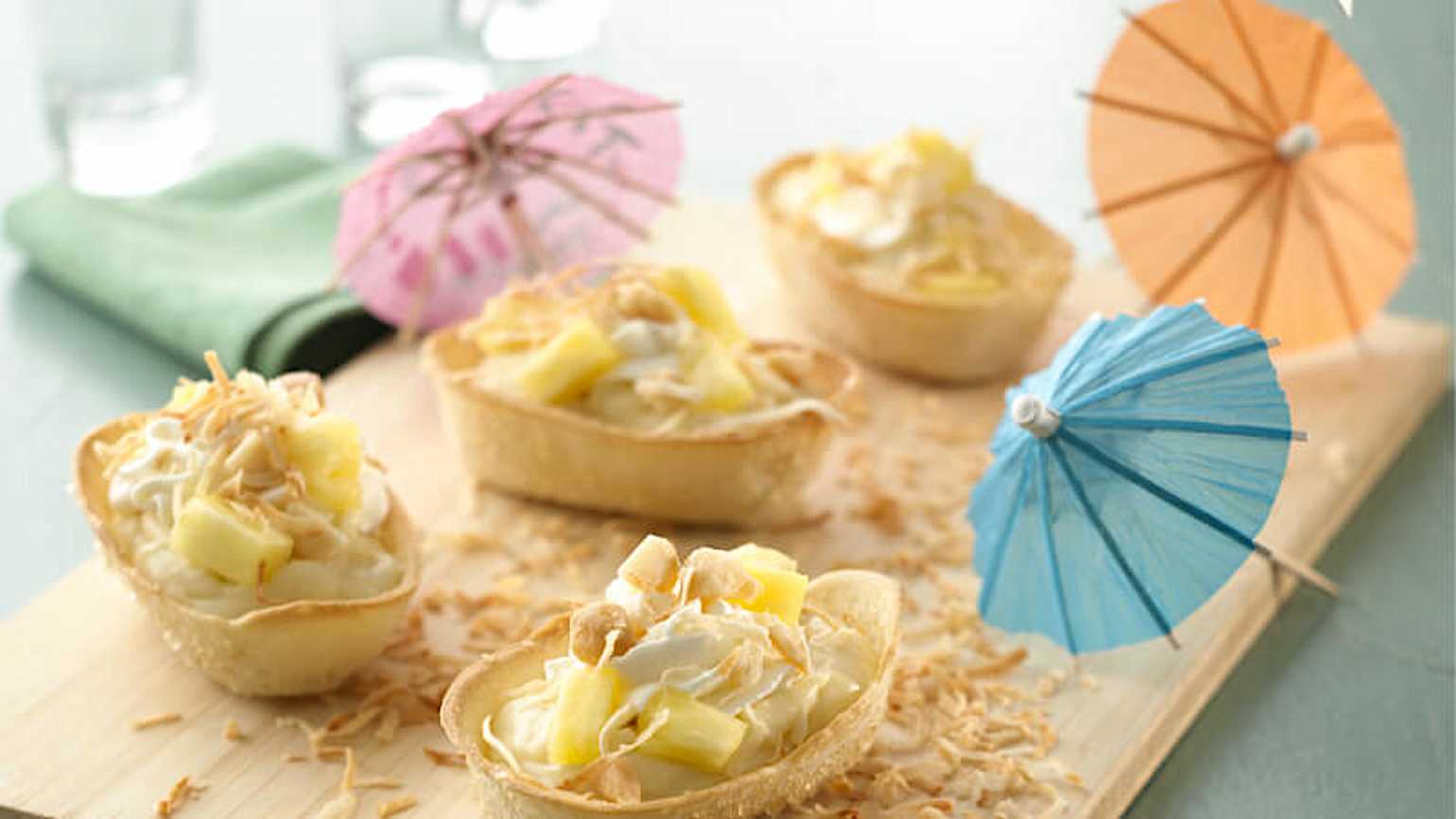 Mini bols à tacos Old El Paso garnis de crème de noix de coco, de morceaux d'ananas et de noix de coco grillée râpée sur une planche de service en bois et de mini parapluies colorés